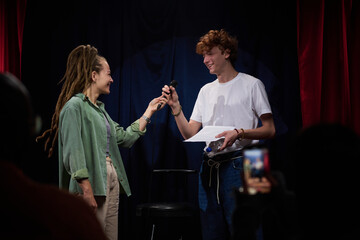 Two people engaging in an interaction on stage, with one person handing a microphone to another who...