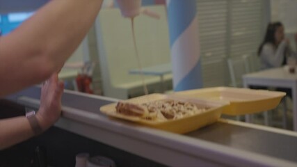A person is pouring a sauce over a pastry in a yellow container. The scene is set in a restaurant or cafe, with a dining table visible in the background