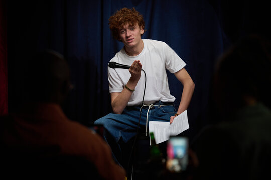 Young man seated on stage holding microphone while interacting with audience during live performance, captured with dramatic lighting setup