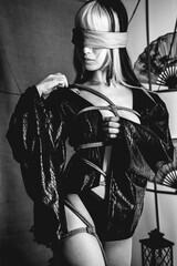 Sexy and beautiful young woman with blonde hair, kimono and panties, and shibari ties and ropes, blindfold, holding a paper fan in a warm japanese room (in black and white)