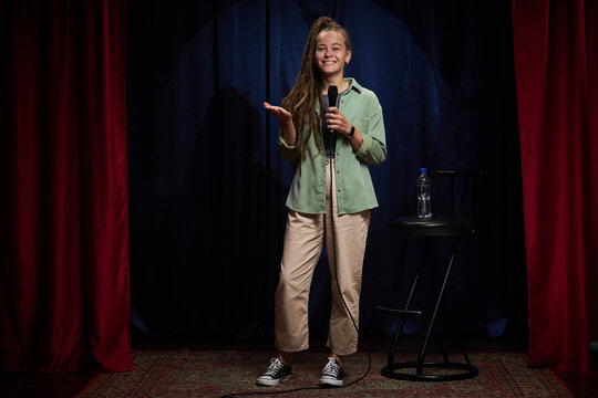 Portrait of smiling girl holding microphone and giving stand-up comedy performance on stage in front of audience wearing casual attire in a spotlight