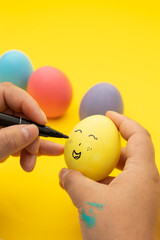 Vertical image of female hands drawing with a marker a happy face on a yellow Easter egg with colored eggs on a yellow background. Easter crafts concept