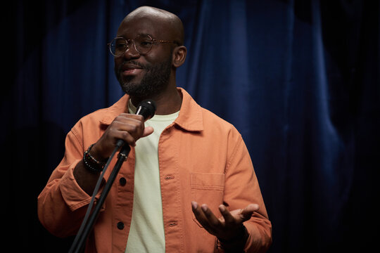 African American man smiling while holding microphone on stage. Wearing glasses and an orange jacket, he appears confident and engaging with audience