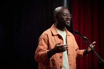 African American man smiling while speaking on microphone during event. Man is wearing orange jacket and standing in front of red and black background