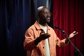 Black person gesturing with hands while speaking into microphone on stage during formal event. Background featuring blue and red curtains