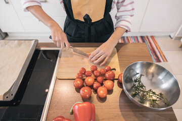 Unrecognizable girl wearing apron slicing fresh tomatoes on wooden cutting board, preparing healthy meal in modern kitchen