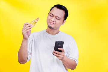 An asian man enjoys a slice of pizza while looking at his smartphone against a yellow background. international foods day concept