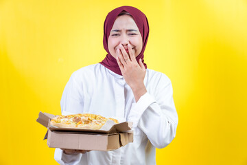 An asian woman with a hijab joyfully reacts to delicious pizza against a bright yellow background.