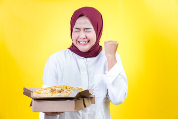 Happy asian woman in hijab celebrates with pizza against a vibrant yellow background
