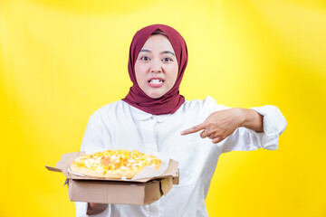 Excited asian woman in a hijab showcasing and pointing delicious pizza against a bright yellow background