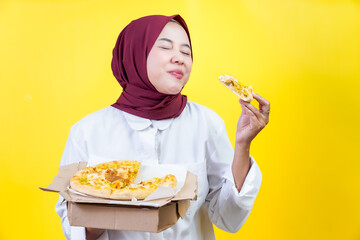 An asian joyful woman in a hijab enjoys pizza against a vibrant yellow background