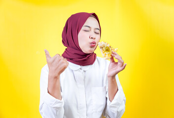 An asian woman in a hijab gives a thumbs up while enjoying slice pizza against a yellow background.