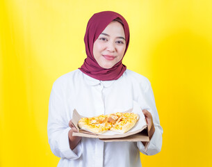 A cheerful asian woman in a hijab holds and showcasing a pizza box against a bright yellow backdrop