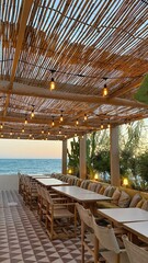 An inviting and romantic outdoor dining area by the sea, featuring a bamboo-covered pergola, string lights and cozy seating with cushions, offering a relaxing afternoon coastal ambiance