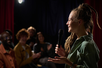 Person delivering stand-up comedy performance on stage with microphone, audience reacting in background, person smiling, emphasizing atmosphere of enjoyment and laughter