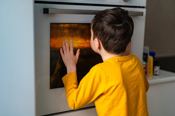 Fototapeta premium A cute boy in a yellow blouse looks into the oven where an apple pie is being prepared. Homemade cakes. The comfort of home. Happy childhood