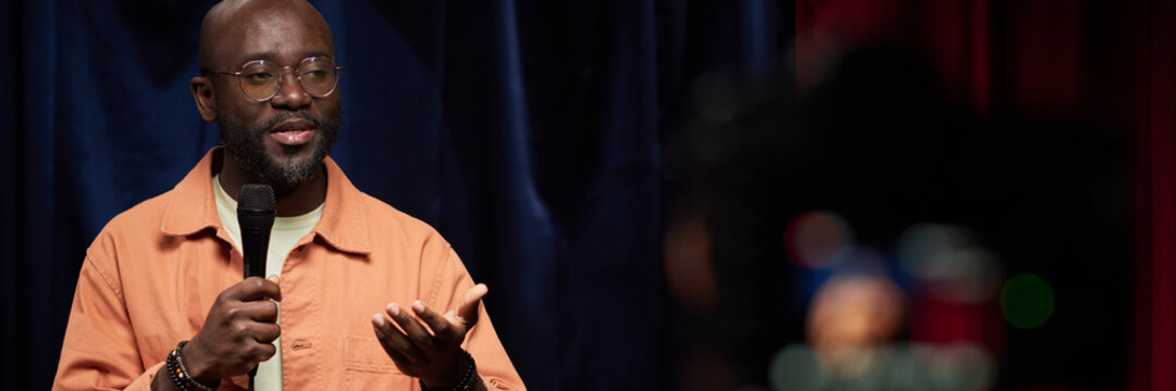 African American man wearing glasses holding microphone while delivering speech on stage bathed in soft light. Background features dark colored curtains enhancing focus on speaker