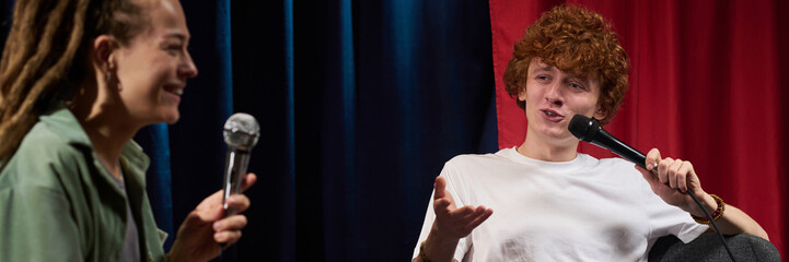Two people engaging in a lively conversation on stage while holding microphones. The setting features vibrant red and blue stage curtains, adding contrast to the scene