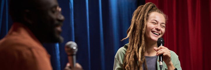 Young woman with dreadlocks holding microphone and smiling, sharing moment of joy. Engaging with audience alongside another individual in vibrant setting