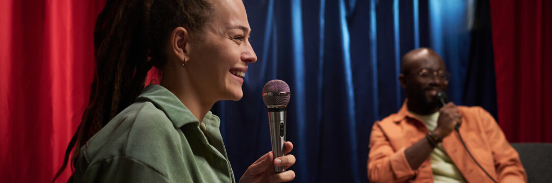 Two individuals actively participating in a talk show recording, with one person speaking into a microphone. Background features curtains in red and blue hues