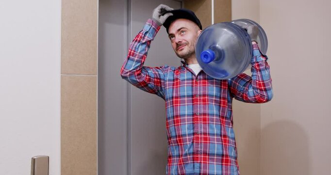 Delivery man with cooler water bottle on shoulder waits for lift. Cheerful man gives thumbs up preparing to deliver container to customer