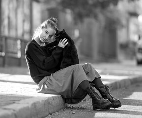 Pretty girl in red skirt hugging black cat outdoors at street with autumn leaves. Beautiful model teenager sitting with feline animal at park