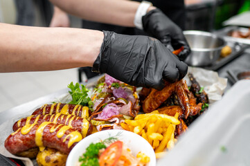 A close-up of a hand wearing a black glove carefully plating a delicious assortment of grilled meats, crispy fries, and fresh vegetables, highlighting vibrant colors and textures in a kitchen setting.