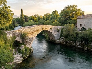 Fototapeta premium Old stone arch bridge over calm river at sunset, lush green trees and plants surround.