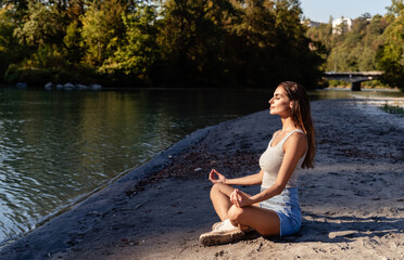 Young woman enjoying sun with closed eyes sitting in lotus position on sand near river