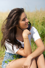Naklejka premium beautiful woman with long brown hair join the summer sun, hold sunglasses, looking to the side, outside in a meadow