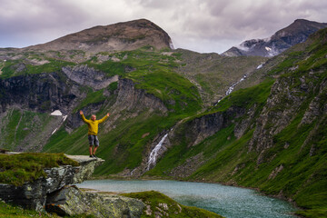 Naklejka premium Happy man standing on rock, hands up and admiring majestic mountain view on famous Grossglockner Hochalpenstrasse road in Austria. Concept: freedom, adventure, harmony with nature