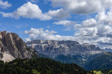 Fototapeta premium The towering cliffs of the Dolomites in Italy rise above dense green forests and rolling valleys, with dynamic cloud formations adding depth to the bright alpine scenery