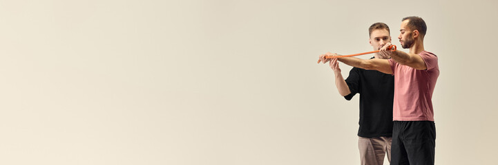 Trainer guiding man in resistance band shoulder exercise, ensuring proper movement for post-training recovery against white studio background. Concept of rehabilitation, recovery, healthcare. Banner