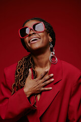 Laughing African American woman with stylish red sunglasses and large hoop earrings against dark red background celebrating and enjoying joyful moment
