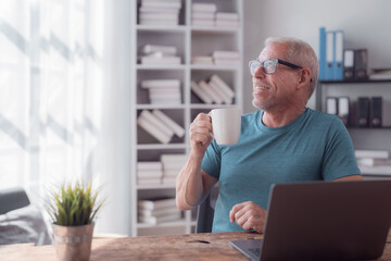Happy retired man drinking coffee and looking outside the window while working with his laptop from his home office, he is enjoying his smart working activity