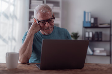 Happy retired man smiling while using a laptop at home, connecting with family online. Embracing modern technology, he enjoys a cozy and relaxed lifestyle during the daytime