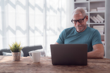 Fototapeta premium Happy retired man working remotely on a laptop at home, enjoying a cup of coffee while surrounded by a plant on a wooden table in a bright and cozy living room