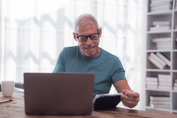 Fototapeta premium Happy retired man working from home using laptop and reading notes from his agenda, sitting at desk in living room with bookcase in background, enjoying his free time and managing his finances