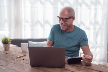 Fototapeta premium Smiling senior man sitting comfortably at a wooden table at home, using a laptop and calculator for managing domestic finances, feeling satisfied with his budgeting and planning