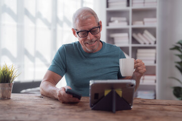 Happy retired man using digital tablet and smartphone while holding a cup of coffee, sitting at wooden table at home, enjoying his free time