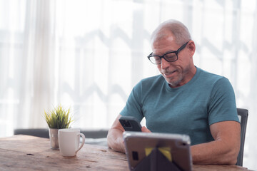 Smiling mature man with eyeglasses using mobile phone and tablet while sitting at wooden table with cup of coffee or tea and small plant in home office, bright window light in background