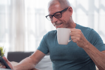 Happy retired man savoring morning coffee while reading news on his smartphone, embracing modern technology and enjoying a connected lifestyle in the comfort of his home