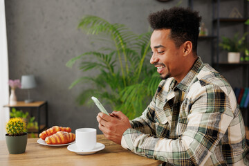 Side view young man of African American ethnicity wears grey casual clothes hold in hand use mobile cell phone sit alone at table in coffee shop cafe relax rest in restaurant during free time indoors.