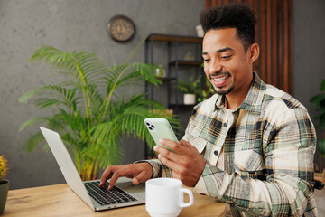 Young smart happy IT man he wears casual clothes glasses hold work on laptop pc computer use mobile cell phone sit alone at table in coffee shop cafe relax rest in restaurant during free time indoors