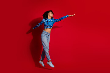 Happy young woman in casual attire posing with a cheerful gesture against a vibrant red background, conveying joy and style.