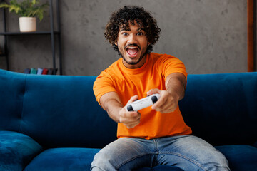 Young Indian man wear orange casual clothes sits on blue sofa couch hold play pc game with joystick...