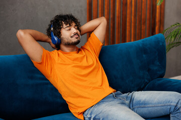 Young happy Indian man wear orange casual clothes sits on blue sofa couch listen to music in...