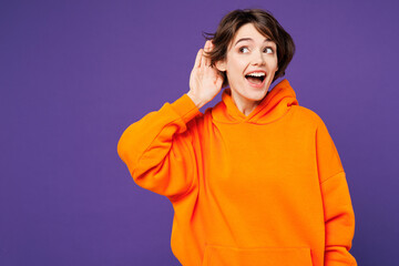 Young smiling curious nosy happy Caucasian woman she wear orange hoody casual clothes try to hear...