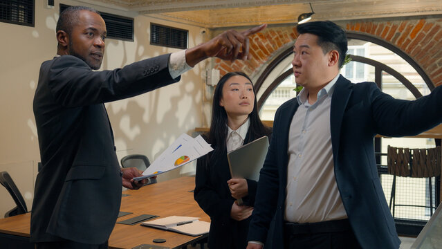 Three multiracial business colleagues coworkers multiethnic in office Asian woman businesswoman Korean Japanese Chinese men businessmen African boss leader talk document discuss pointing data diagram