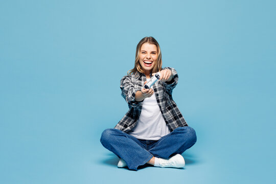 Full body young woman she wears checkered grey shirt casual clothes sit hold in hand play pc game with joystick console isolated on plain pastel light blue cyan background studio. Lifestyle concept.